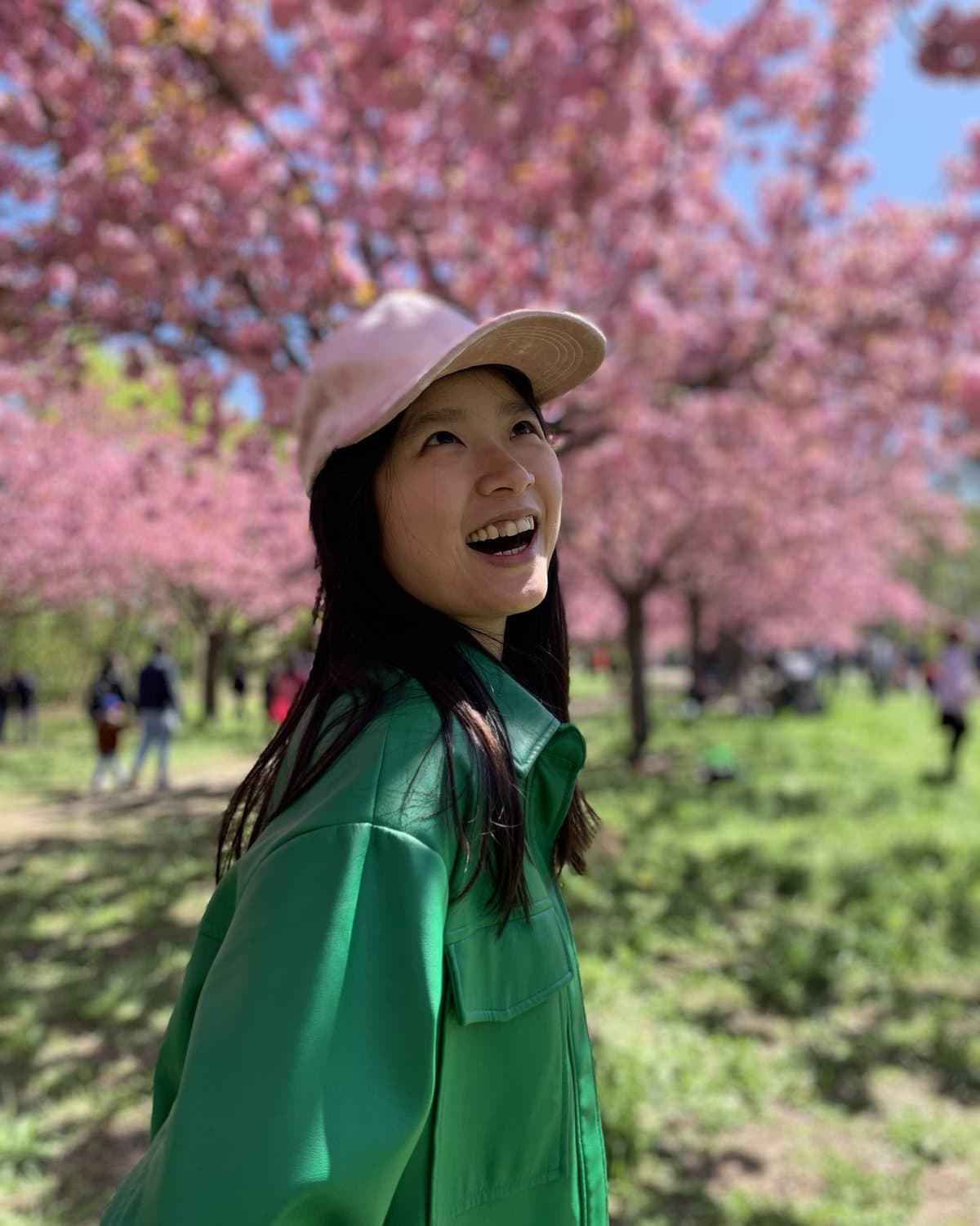 Shirley Xue laughing under cherry blossoms in Berlin — pink cap, green jacket